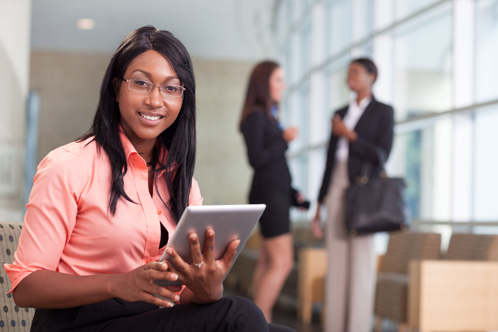 african-american business woman sitting in lobby with computer tablet, looking at camera, smiling, two business women in background